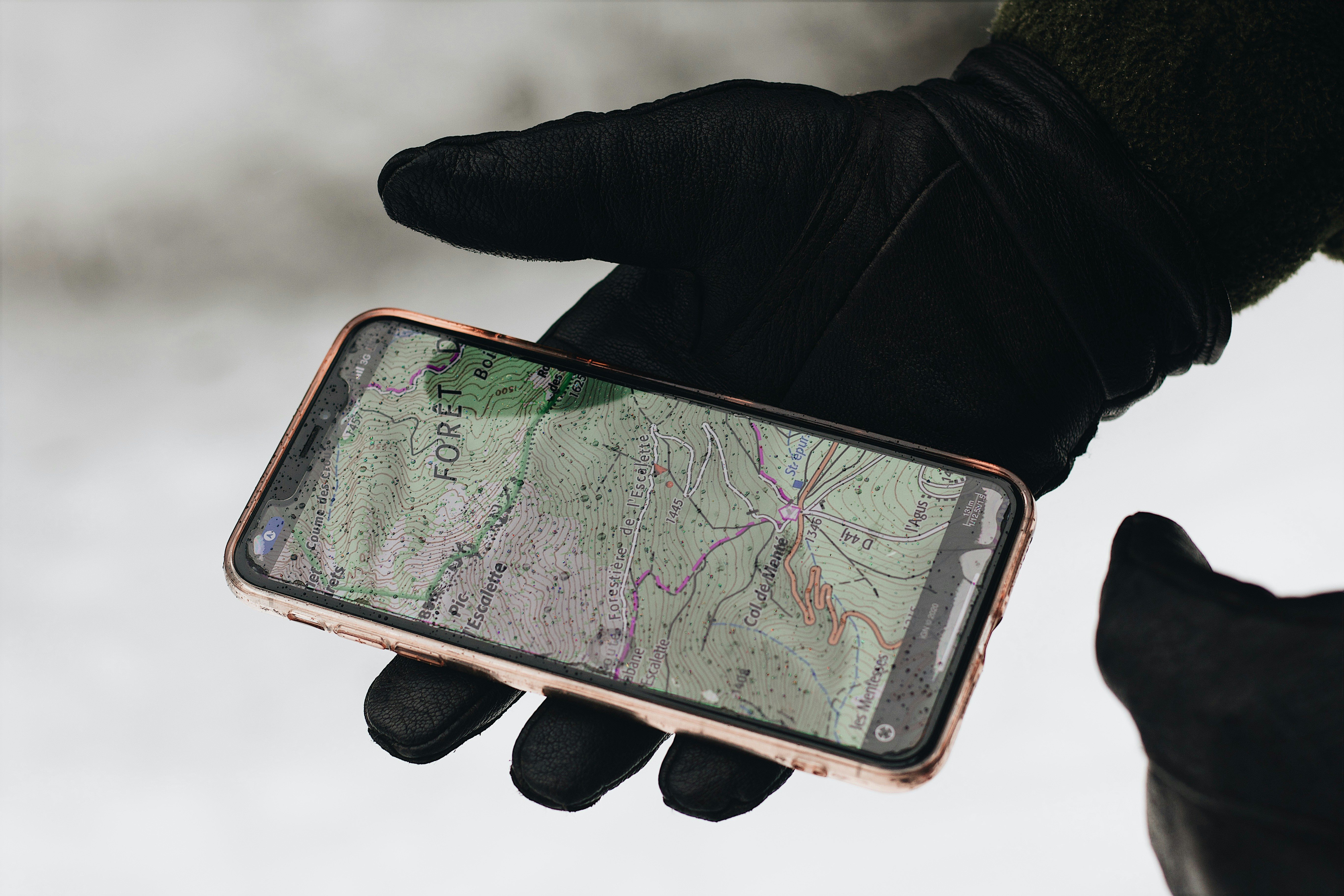 Hand wearing black glove holding a smartphone displaying a topographic map with forest and trail details.